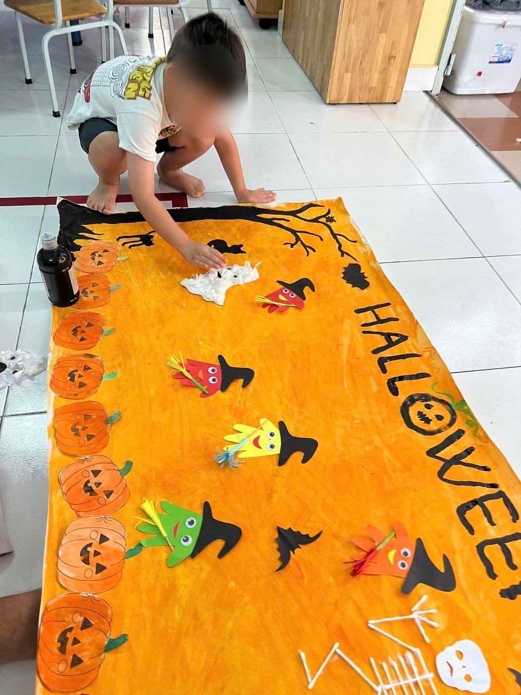 A child sticks decorations onto a large orange Halloween mural laid on the floor
